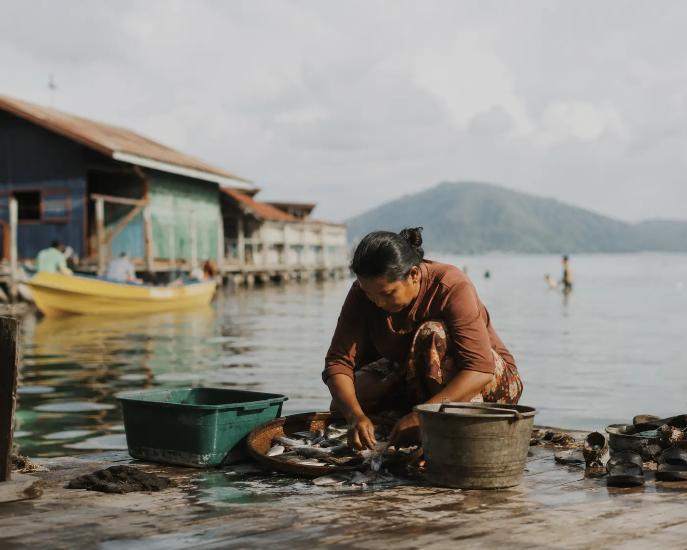A woman sorting the day's catch on a wooden jetty, stilt houses and moored boats behind her.